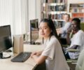 Asian young lady, black young man and white young mand seating in a study room in front of monitors and keyboards